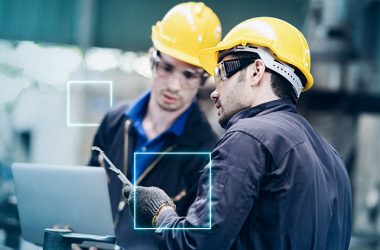 Dois homens com capacete amarelo olham para um computador dentro de uma indústria.