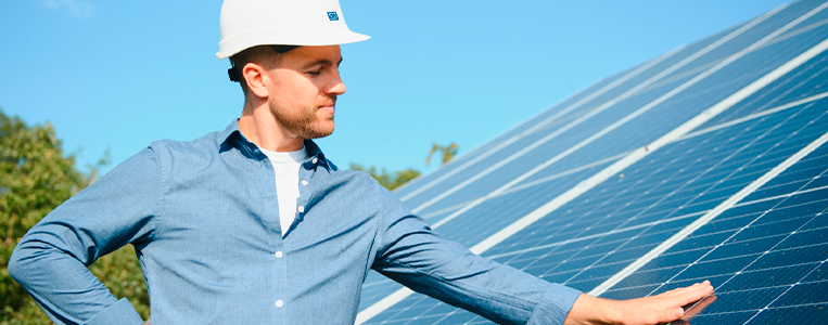Homem analisando painéis solares sob céu claro, usando capacete branco.