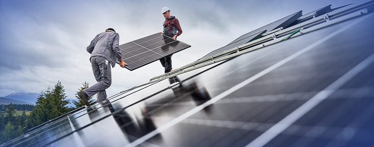 Trabalhadores instalando painéis solares em um telhado com o céu ao fundo, promovendo energia renovável e sustentável.