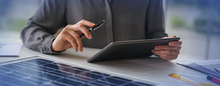 Pessoa em escritório segura um tablet e uma caneta, com um painel solar em primeiro plano sobre a mesa, sugerindo revisão final do projeto.
