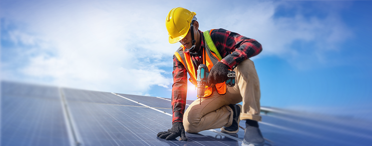 Técnico ajoelhado sobre painéis solares no telhado, usando ferramentas e EPIs (capacete e colete), representando uma instalação que exige cuidado e precisão.


