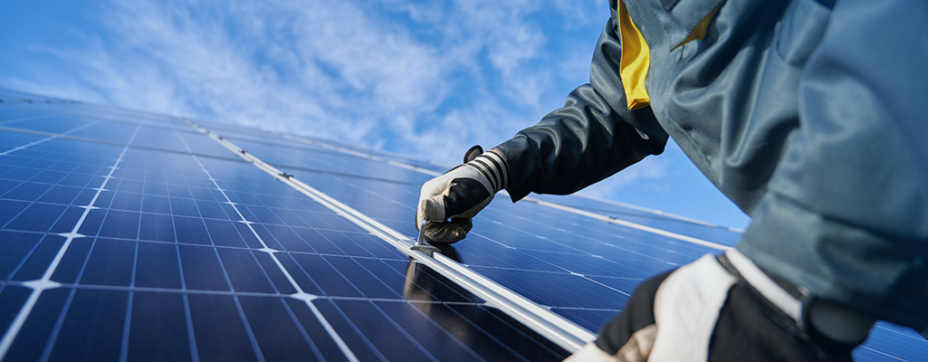Close de mãos com luvas ajustando a estrutura/encaixe entre módulos fotovoltaicos sob céu azul, destacando detalhes técnicos de montagem e manutenção.