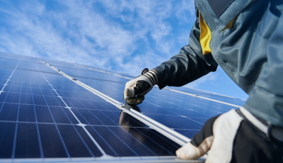 Close de mãos com luvas ajustando a estrutura/encaixe entre módulos fotovoltaicos sob céu azul, destacando detalhes técnicos de montagem e manutenção.