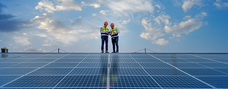 Dois técnicos ao longe, sobre uma grande área de painéis solares, analisam documentos/prancheta sob um céu parcialmente nublado.