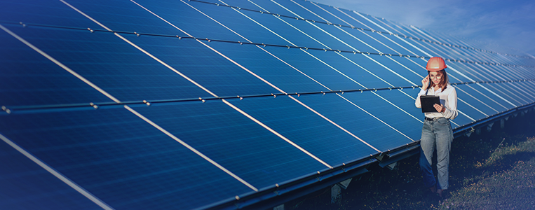 Profissional com capacete e prancheta/tablet caminha ao lado de uma usina solar, inspecionando fileiras de painéis em um ambiente aberto.
