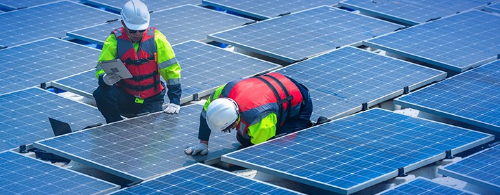 Dois técnicos com capacete e colete trabalham entre painéis solares, um com tablet e outro ajustando um módulo, ilustrando inspeção e etapas de homologação.