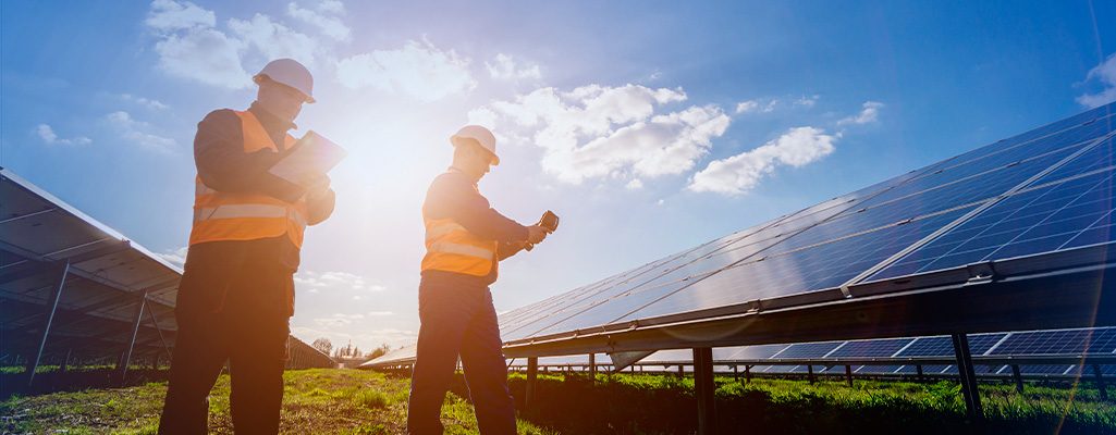 Dois profissionais em uma usina solar analisam prancheta e celular ao lado de painéis, com sol ao fundo, representando vistoria e validação do projeto.