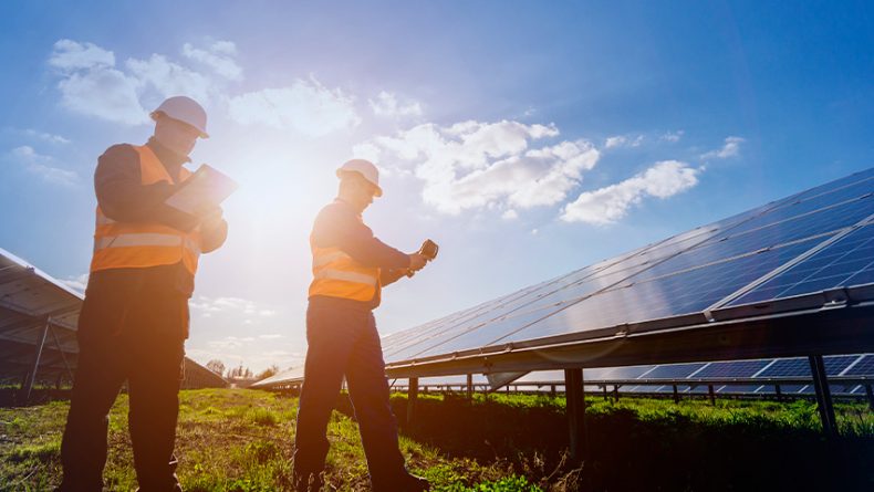 Dois profissionais em uma usina solar analisam prancheta e celular ao lado de painéis, com sol ao fundo, representando vistoria e validação do projeto.
