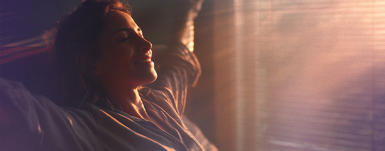 Mulher sorridente apreciando o pôr do sol, relaxando e aproveitando momentos de paz e tranquilidade ao lado da janela