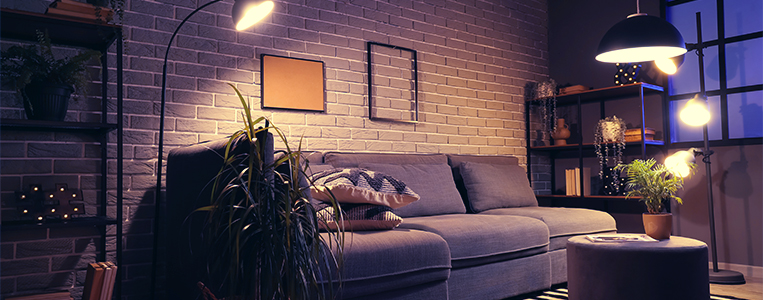 Imagem de uma sala de estar moderna com sofá cinza, iluminação quente, prateleiras com livros, plantas e decoração minimalista com parede de tijolos expostos.