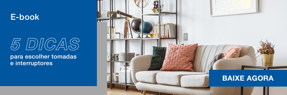 Imagem de uma sala de estar moderna com sofá bege, almofadas coloridas, prateleira de aço com decoração e uma estante de livros, ideal para aprender sobre tomadas e interruptores.