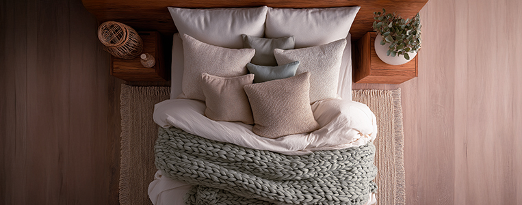 Cama de casal com roupa de cama branca, travesseiros em tons neutros e uma manta de tricô cinza sobre um piso de madeira, decorada com uma planta em um criado-mudo ao lado.