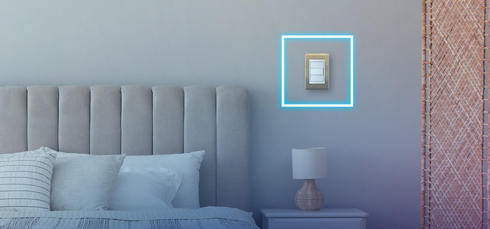 Imagem de um interruptor de luz moderno instalado na parede de um quarto bem decorado, com cabeceira de cama acolchoada e luminária de mesa ao lado.