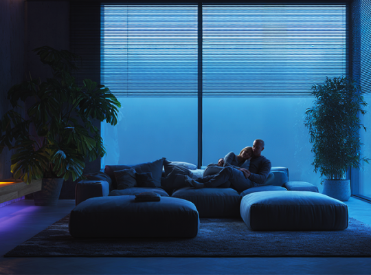 Sala de estar moderna com iluminação azul suave e plantas, onde um casal relaxa em um sofá grande diante de janelas amplas.