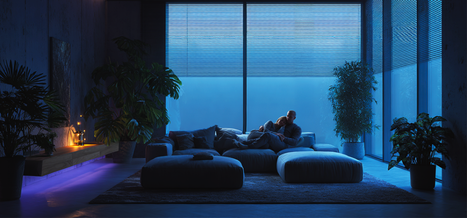 Sala de estar moderna com iluminação azul suave e plantas, onde um casal relaxa em um sofá grande diante de janelas amplas.