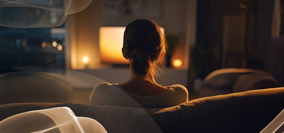 Pessoa vista de costas em uma sala aconchegante com iluminação baixa e TV acesa, com elementos gráficos suaves sobrepostos, sugerindo experiências conectadas e automação no lar.