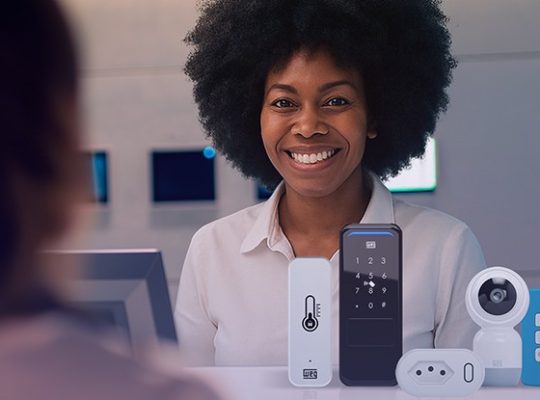 Atendente sorrindo atrás do balcão com vários dispositivos de automação residencial expostos (teclado, câmera e módulos), sugerindo atendimento e demonstração de produtos.