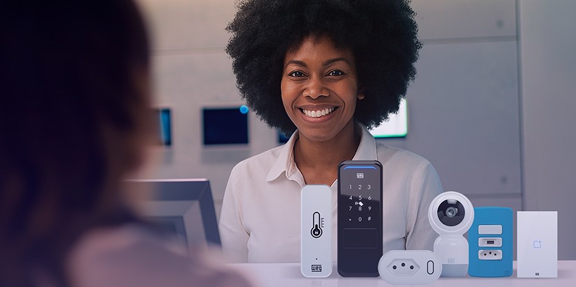 Atendente sorrindo atrás do balcão com vários dispositivos de automação residencial expostos (teclado, câmera e módulos), sugerindo atendimento e demonstração de produtos.