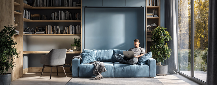 Sala integrada com estante e mesa de trabalho; pessoa usando notebook no sofá, indicando um espaço que mistura lazer e home office.