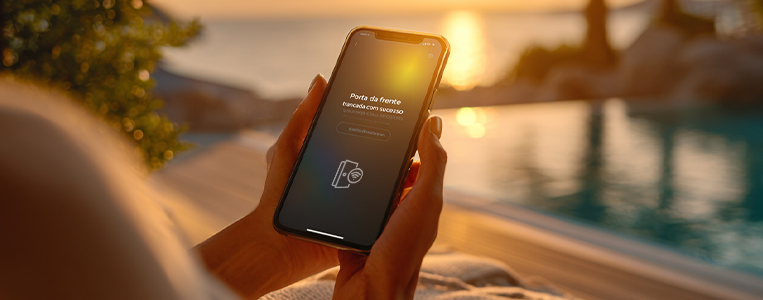 Pessoa segura um smartphone exibindo aviso de “Porta da frente”/segurança, com piscina e pôr do sol ao fundo, sugerindo monitoramento remoto.