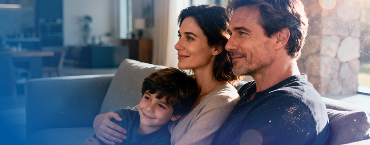 Família no sofá (duas pessoas adultas e uma criança) em ambiente aconchegante, transmitindo conforto e satisfação com a casa conectada.