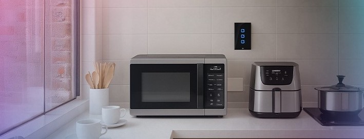Bancada de cozinha com micro-ondas, air fryer e panela; ao fundo, um interruptor/painel inteligente na parede, indicando controle de dispositivos.